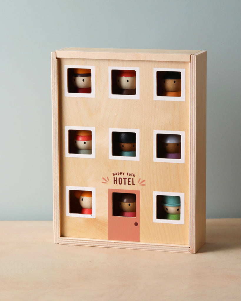 A solid wood toy shaped like a building named "Happy Folk Hotel" with nine compartments, each containing a peg doll head. The peg dolls, wearing different colored hats, can be seen through the square windows. Perfect for open-ended play, it features a door with a doll visible behind it.