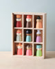 A solid wood display box with nine compartments, each containing a small, colorful wooden figure with simplistic features and various headgear. Perfect for open-ended play, the Happy Folk Hotel features figures painted in vibrant colors like red, green, purple, and blue and have a minimalist design to inspire creative play.