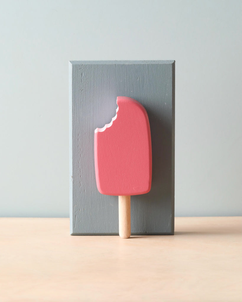 Wooden handmade bright pink popsicle (raspberry flavor). Leaning against light blue wooden block. 