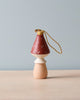 The Handmade Wooden Mushroom Ornament, measuring 4 inches tall, features a beige body and a red cap adorned with white polka dots, topped with a golden string loop for hanging. It is displayed on a light-colored surface against a pale blue background.