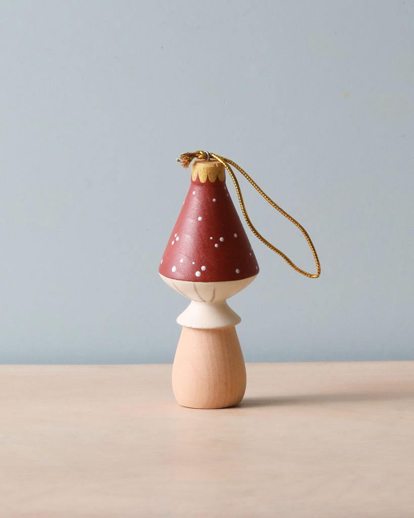 The Handmade Wooden Mushroom Ornament, measuring 4 inches tall, features a beige body and a red cap adorned with white polka dots, topped with a golden string loop for hanging. It is displayed on a light-colored surface against a pale blue background.