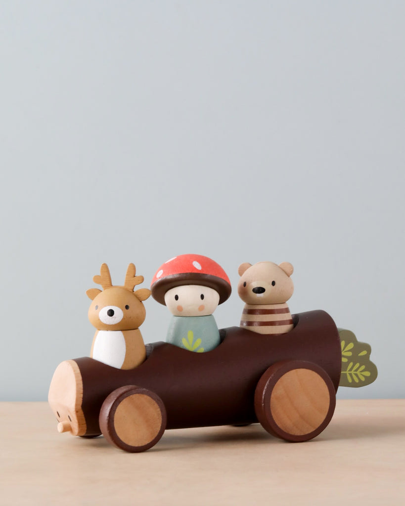 A Timber Taxi, painted brown with leaf details, holds three Merrywood folks: a person in a red hat, and two animals, a deer and a bear, against a plain, light blue