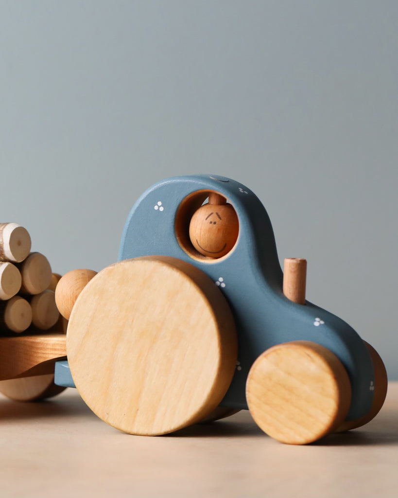 A Handmade Wooden Tractor painted in blue pulls a detachable trailer filled with small cylindrical logs. The tractor features large circular wheels and a small wooden driver in the cab, smiling brightly. The background is plain, with a light neutral color.