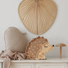 A cozy, neutral-colored setup featuring a Little Lights Hedgehog Lamp on a white surface. The scene includes a leaf-shaped cushion, a soft beige blanket, a Little Lights Hedgehog Lamp shaped like a mushroom, and a large fan-shaped decorative piece hanging on the wall. Perfect for a children's night light corner.