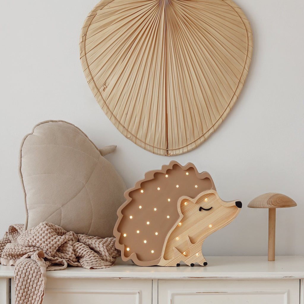 A cozy, neutral-colored setup featuring a Little Lights Hedgehog Lamp on a white surface. The scene includes a leaf-shaped cushion, a soft beige blanket, a Little Lights Hedgehog Lamp shaped like a mushroom, and a large fan-shaped decorative piece hanging on the wall. Perfect for a children's night light corner.