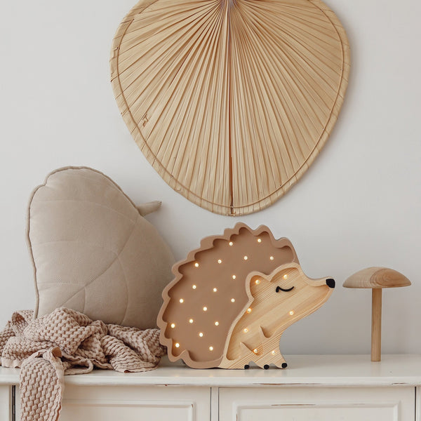 A cozy, neutral-colored setup featuring a Little Lights Hedgehog Lamp on a white surface. The scene includes a leaf-shaped cushion, a soft beige blanket, a Little Lights Hedgehog Lamp shaped like a mushroom, and a large fan-shaped decorative piece hanging on the wall. Perfect for a children's night light corner.