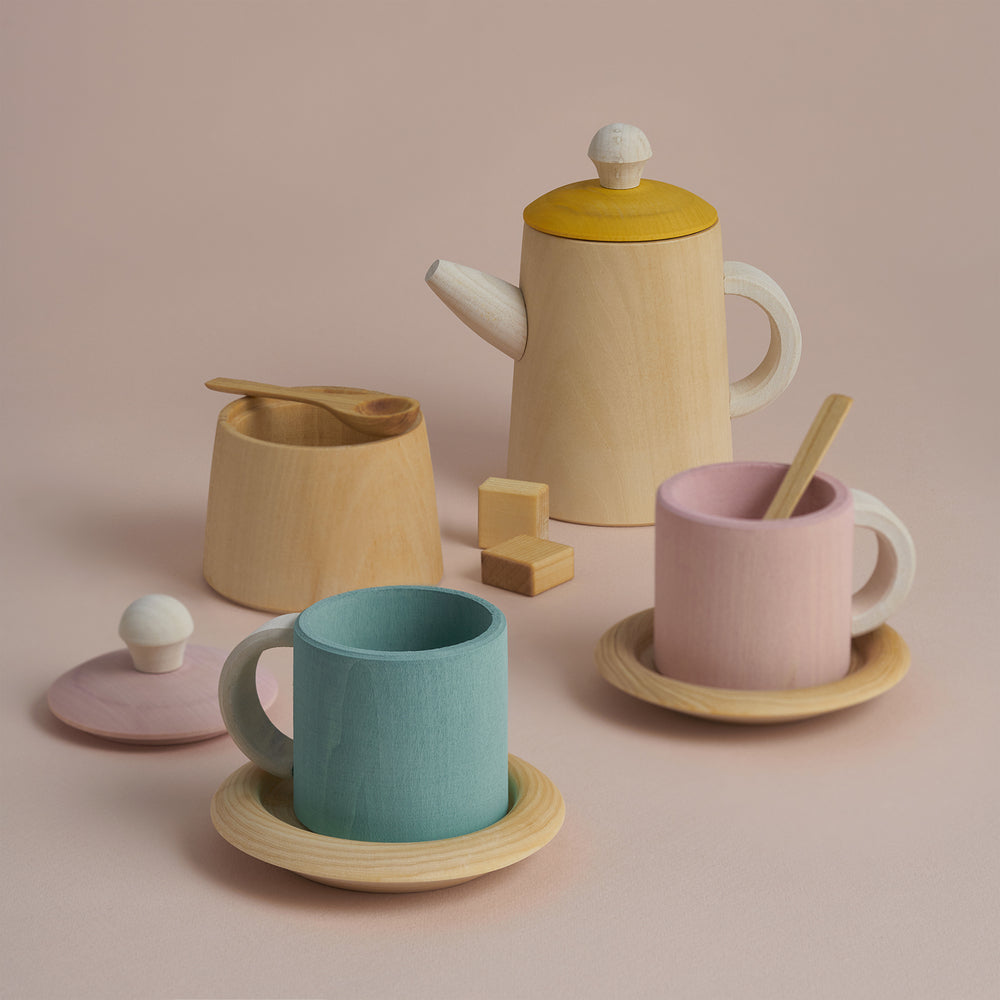 A handmade pastel-colored "Raduga Grez Handmade Wooden Tea Set" on a light pink background. The set, painted with non-toxic paint, includes a wooden teapot with a yellow lid, a sugar bowl with a spoon, a blue cup on a saucer, and a pink cup with a stirrer on its own saucer. Perfect for pretend play!