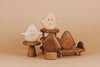 A collection of Wooden Ghost Emotions, each displaying basic emotions with a unique, simple expression, is arranged on various wooden stands and chairs against a beige background. The figures are made from different shades of wood and finished with natural linseed oil, giving them a handcrafted appearance.