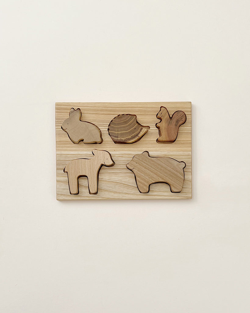 A Handmade Wooden Woodland Puzzle featuring cut-out animal shapes, including a bear, squirrel, pig, snail, and another bear. The light-colored wooden pieces are set against a matching background and treated with beeswax for a smooth finish, all beautifully mounted on a wall.