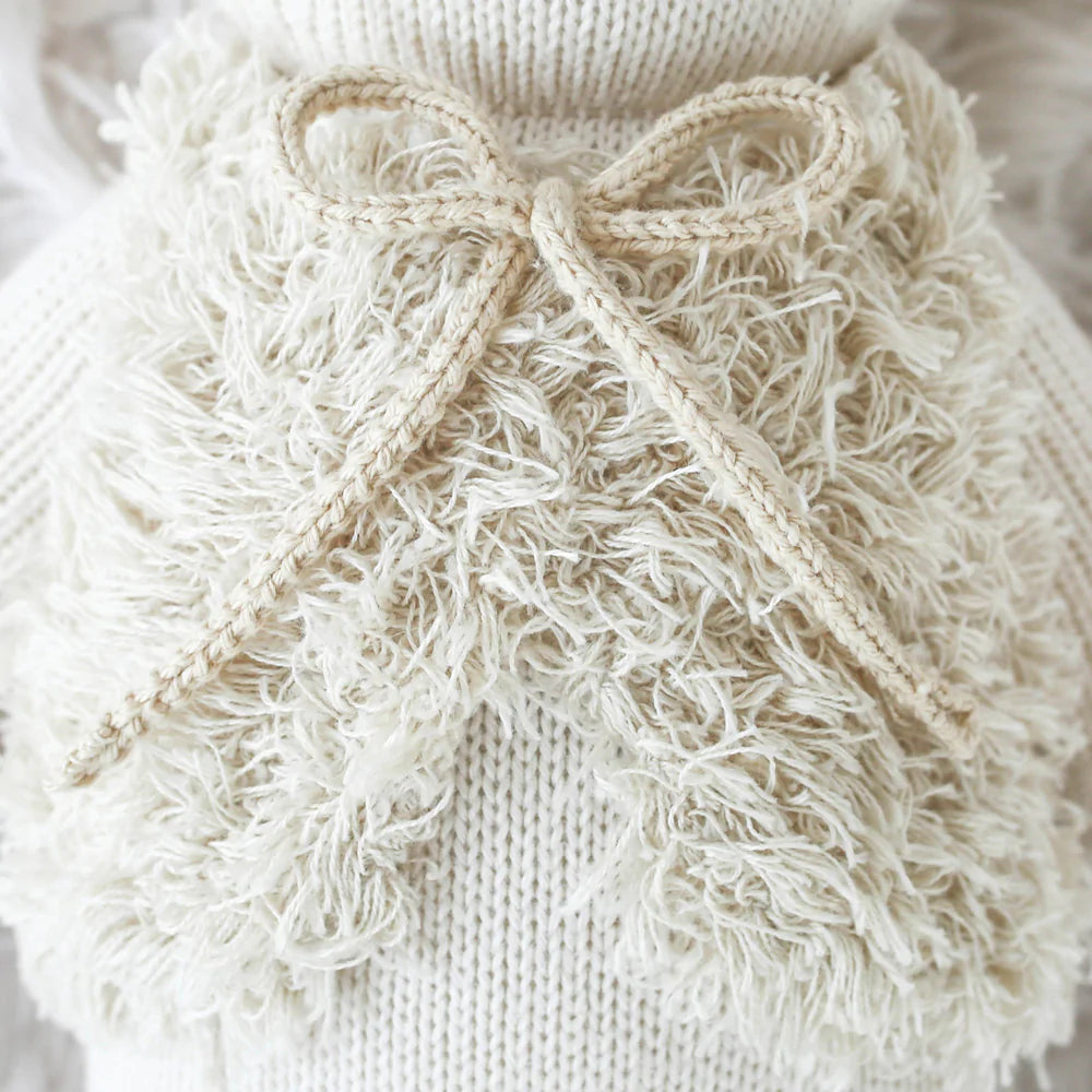 Close-up image of the hand-knit Cuddle + Kind Stella The Polar Bear, showcasing its cream-colored material. The bear features a thick, textured fluffy section in front and a decorative bow made from the same yarn tied neatly at the center. Made with hypoallergenic fibers and adhering to fair trade practices, against a similarly cream-colored background.