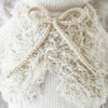 Close-up image of the hand-knit Cuddle + Kind Stella The Polar Bear, showcasing its cream-colored material. The bear features a thick, textured fluffy section in front and a decorative bow made from the same yarn tied neatly at the center. Made with hypoallergenic fibers and adhering to fair trade practices, against a similarly cream-colored background.