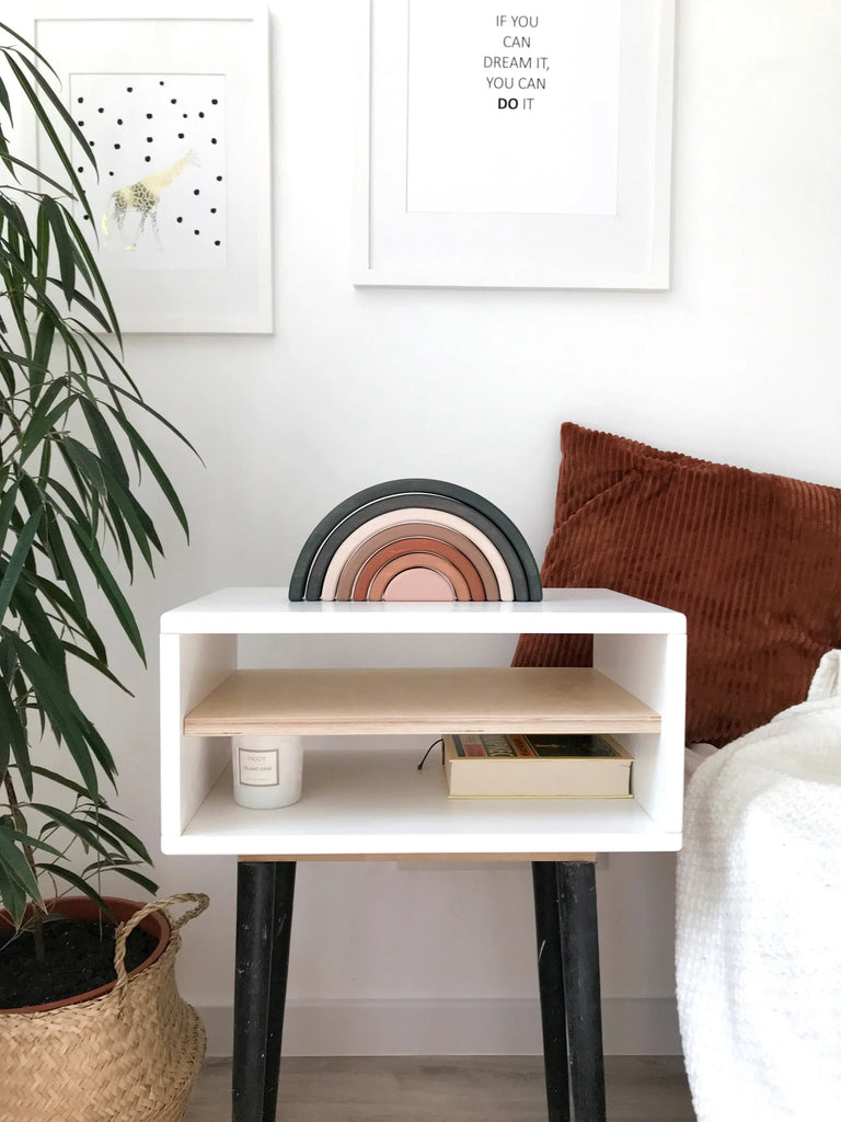 A minimalist side table with a modern white shelf and black legs. The shelf holds a candle, a book, and the Handmade Rainbow Stacker - Mojave Desert, painted with non-toxic paint. A textured cushion is nearby, and two framed prints hang on the wall; one includes a motivational quote. A plant in a basket sits beside the table.