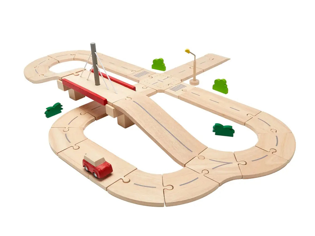 Introducing the Wooden Road System Deluxe: a wooden toy road set with interlocking pieces forming a figure-eight layout. It includes a bridge, a small red toy car, a lamppost, and green tree figures. Sustainably made for pretend-play transportation, the road pieces feature printed lines and markings to simulate real roadways.