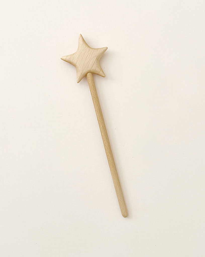 A Handmade Wooden Magic Wand, carved from beech wood, lies flat on a light beige surface. The wand features a simple design with a straight handle and a star-shaped top, showcasing its natural light-colored wood.