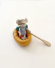 A small mouse figurine wearing a striped shirt and a sun hat sits in a Maileg Rubber Boat. It holds a wooden paddle, ready to row on its pretend water adventure. The mouse is surrounded by minimal white background, highlighting the whimsical scene.