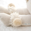 Close-up of a knitted soft toy featuring cream-colored yarn and hypoallergenic materials. The focus is on its fluffy, pom-pom adorned feet. A "cuddle + kind" label is partially visible, suggesting the fair trade brand of the hand-knit toy. The background is a soft, textured white fabric. This adorable toy is known as Cuddle + Kind Stella The Polar Bear.