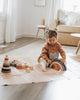 A young child sits on a mat in a bright, airy room, playing with handmade wooden blocks and the Handmade Rainbow Stacker - Mojave Desert painted with non-toxic paint. The child is wearing a brown sweater and has a bow in their hair. In the background, there is a beige sofa with cushions and a small table with decorative items.