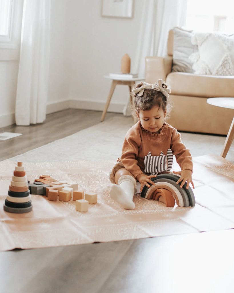 A young child sits on a mat in a bright, airy room, playing with handmade wooden blocks and the Handmade Rainbow Stacker - Mojave Desert painted with non-toxic paint. The child is wearing a brown sweater and has a bow in their hair. In the background, there is a beige sofa with cushions and a small table with decorative items.