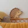 A cozy bedroom features the Little Lights Hedgehog Lamp with small illuminated spots, mounted on a white wall. Below is a woven rattan furniture piece with yellow and beige cushions, adding a warm and inviting atmosphere to the room. This adorable children's night light adds charm and comfort.