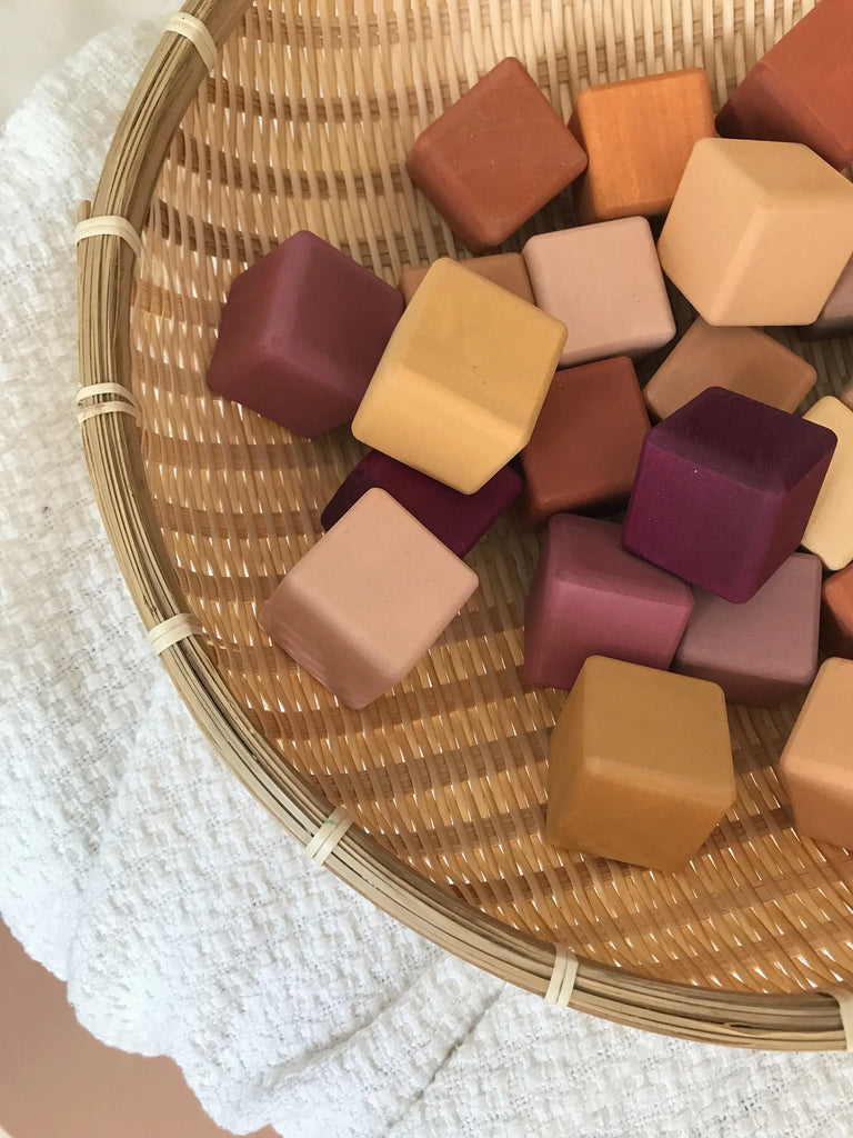 A wicker basket filled with colorful square and hexagonal wooden blocks in shades of red, purple, and beige, painted with non-toxic paint, displayed on a white textured cloth.