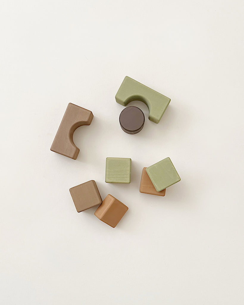 A minimalist set of Handmade Wooden Building Blocks crafted from linden wood, featuring a variety of shapes such as rectangles, squares, a circle, and a semicircle in earthy brown and green hues against a light backdrop. Ideal for enhancing fine motor skills through creative play.