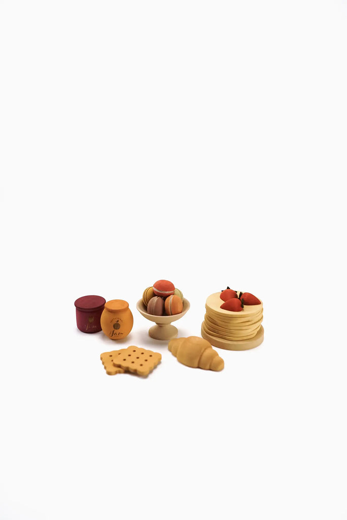 A small arrangement of pretend play food on a white background includes a croissant, crackers, macarons, a jar of honey, and a jar of jam, along with a stack of pancakes topped with strawberries. This delightful Sabo Concept Handmade Wooden Bakery Set is painted with non-toxic paint for safe and imaginative play.