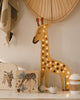 In a cozy nursery setup, you can find the Little Lights Giraffe Lamp perched on a shelf, emanating a gentle glow as a children's night light. Nearby are books adorned with giraffe illustrations and several small decorative objects. Above it all, a handwoven fan hangs to infuse warmth that complements the charm of the natural pinewood.