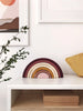 A white table features the Handmade Rainbow Stacker - Pink, displaying a decorative wooden rainbow in soft shades of pink, yellow, and purple made from non-toxic paint. A green leafy branch extends from the left side. In the background, there is framed wall art, and a yellow cushion is partially visible on the right side.