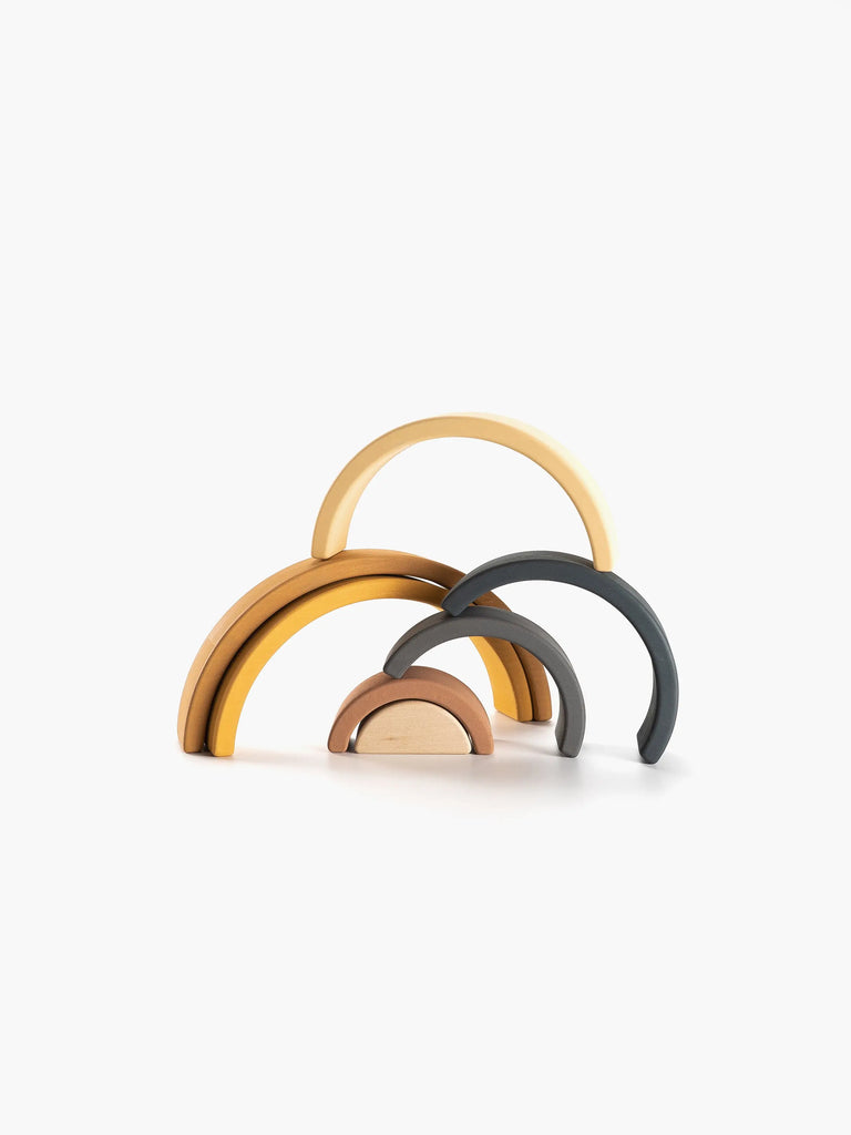 The Handmade Rainbow Stacker - Mustard features a set of interlocking, semi-circular wooden arches in various shades of yellow, brown, and gray, arranged in a minimalistic, abstract formation on a white background. This non-toxic, handmade stacking toy resembles a simplified, stylized rainbow.