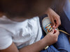 A child wearing a white shirt and jeans sits, skillfully tying the laces of a Tie Up Shoe, which features blue fabric with orange details, showcasing impressive hand-eye coordination.