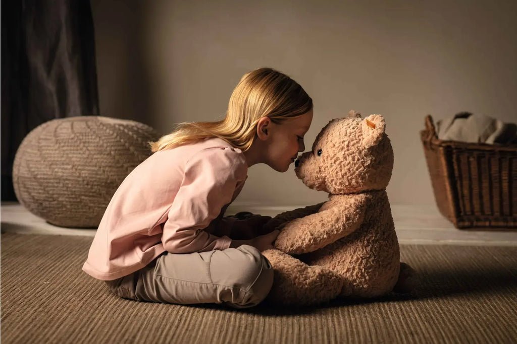 A young girl in a pink shirt gently touches her forehead to the nose of a large, plush Steiff XL Jimmy Teddy Bear, 22 Inches as they sit on a carpeted floor, with soft lighting enhancing the intimate moment.