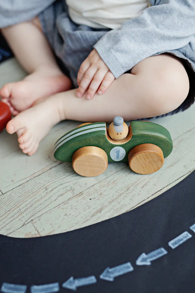 A young child sits on the floor, their legs crossed, playing with a Handmade Wooden Toy Race Car made from sustainably harvested birch wood, adorned with the number "1". The child wears shorts and a gray sweater. The toy car is placed on a track mat with arrows pointing in one direction.