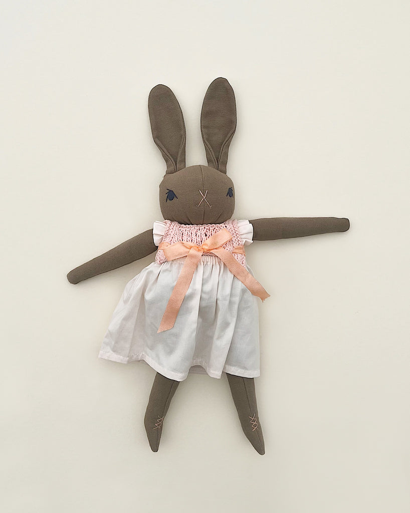 A handmade Polka Dot Club Large Brown Rabbit in Hand Knit Dress with a peach bow and white dress against a light background. The rabbit, hand embroidered in Peru, has long ears, blue stitched eyes, and an X for a mouth.