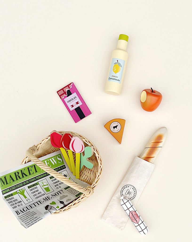 A light, handcrafted shopping basket (ships in approximately one week) holds colorful faux tulips, a toy newspaper, a wrapped toy baguette, and a toy cheese wheel piece. Nearby for pretend play are a toy bottle of lemonade, a toy chocolate bar, and a plastic apple, all arranged on a white surface.