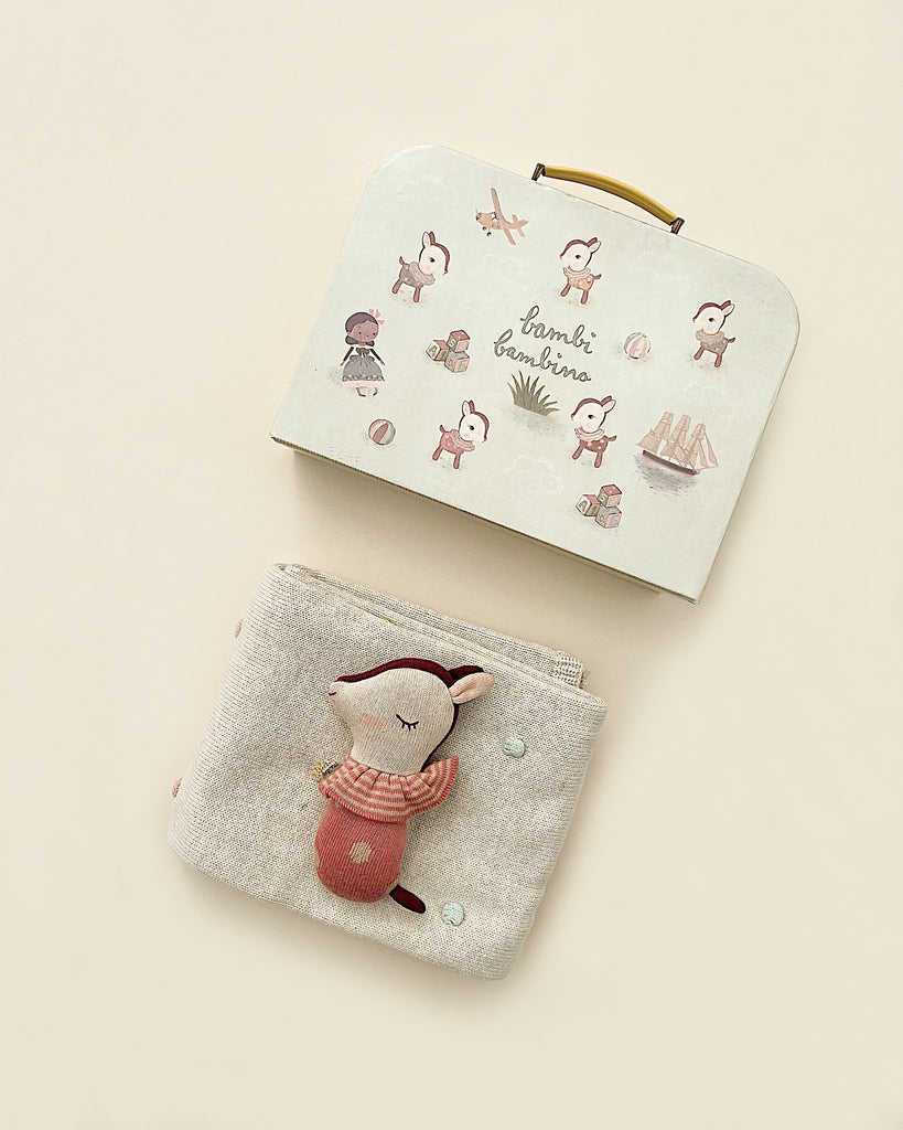 A small suitcase with a handle, decorated with illustrated deer and objects, lies against a beige background. Below it, there is a fabric-covered book featuring a small, soft toy deer wearing a striped scarf and pink pants—perfect for a Maileg Baby Gift Set.