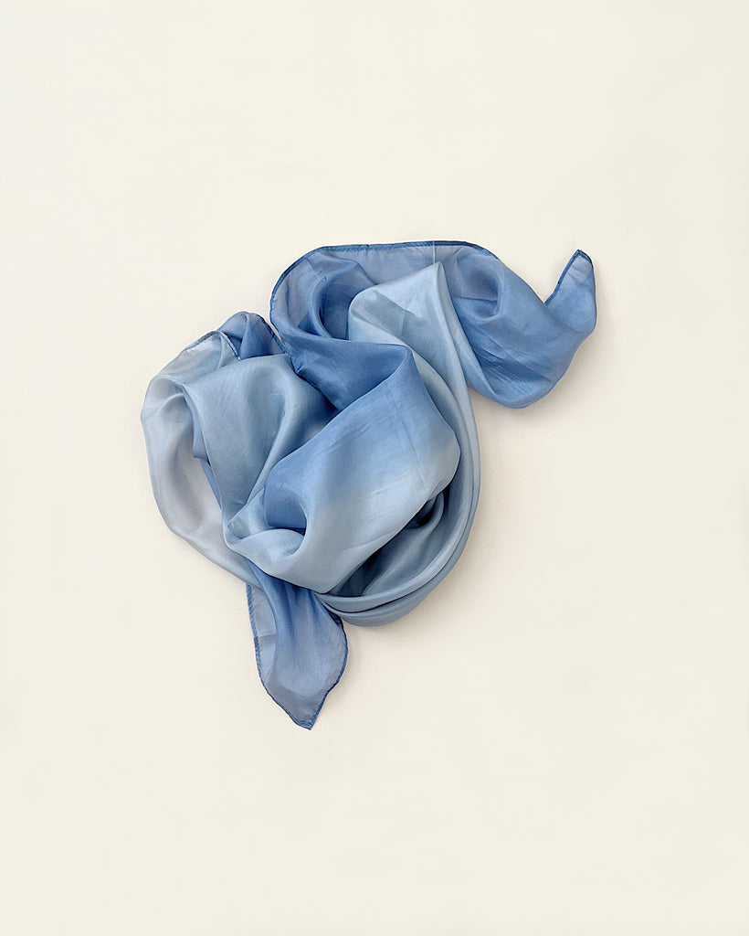 A soft, light blue Sarah's Silk Earth Playsilk - Stone elegantly arranged in a loose, flowing swirl on a plain beige background.