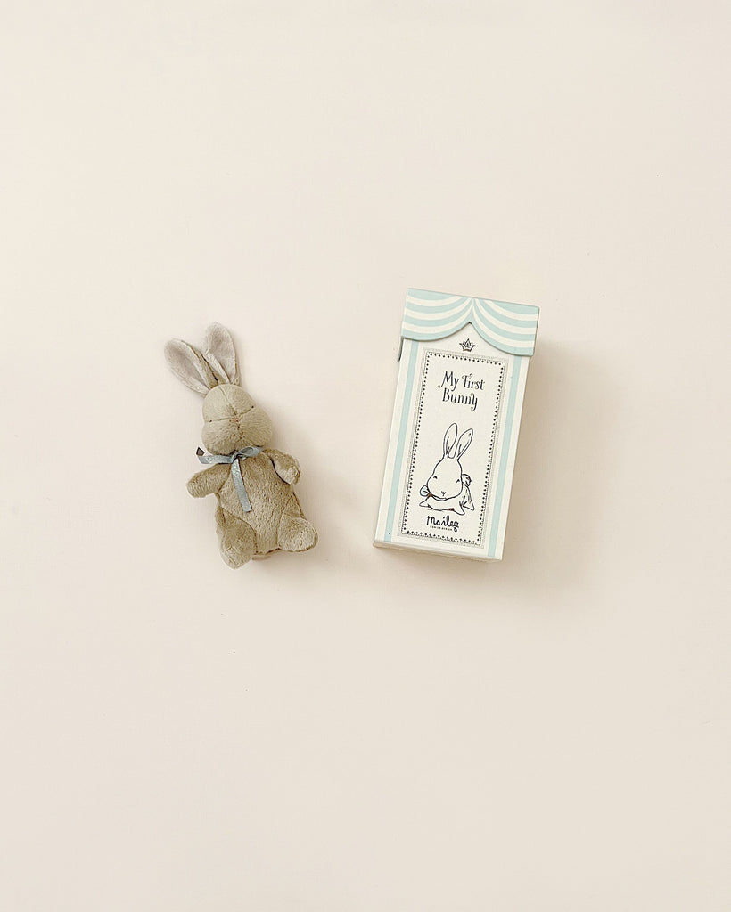 A small, soft plush bunny with a blue ribbon around its neck lies next to a box labeled "Maileg My First Bunny - Light Blue" against a plain white background. The box features an illustration of a bunny and a blue and white striped design, making it an ideal baby shower gift.