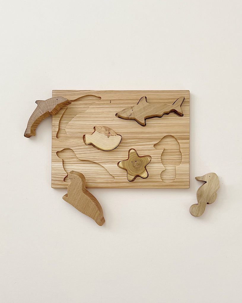 A Handmade Wooden Ocean Puzzle featuring cutouts in the shape of various sea creatures, including a dolphin, fish, starfish, seahorse, and others. Crafted from different kinds of wood and finished with linseed oil and beeswax, some pieces are placed next to the board on a plain white background.