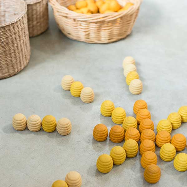 Various Grapat Mandala Honeycomb pieces from sustainable forests in shades of cream and yellow are scattered across a table, with some arranged to spell "hi." A wicker basket filled with similar pieces is in the background.