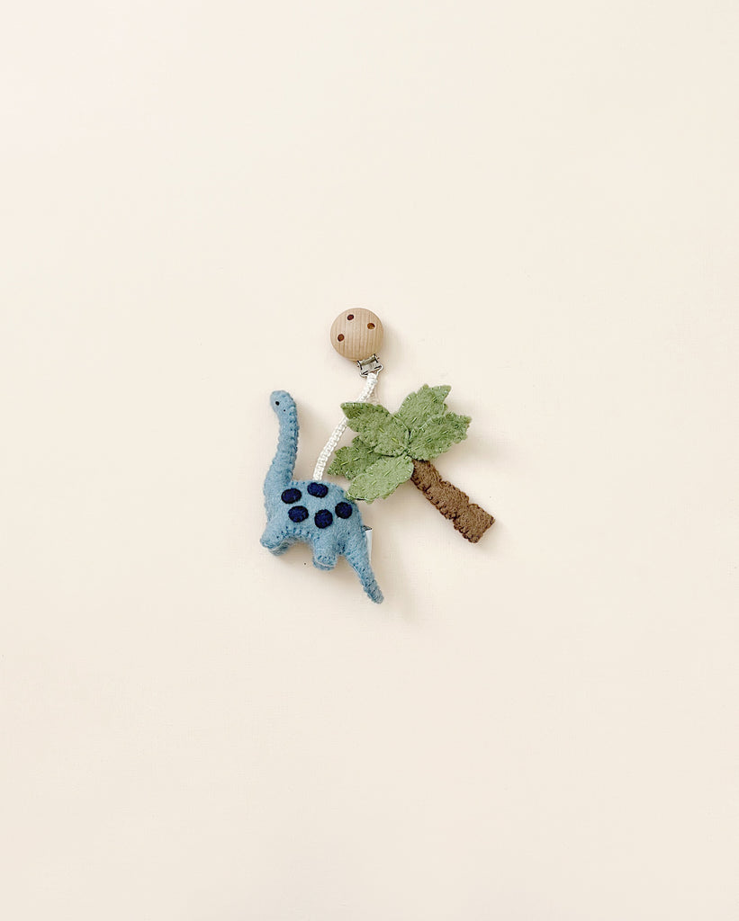 A small, handmade in Nepal Dinosaur Pram Clip with blue wool crochet body and black spots, accompanied by a fabric leaf and twig, against a plain light background.