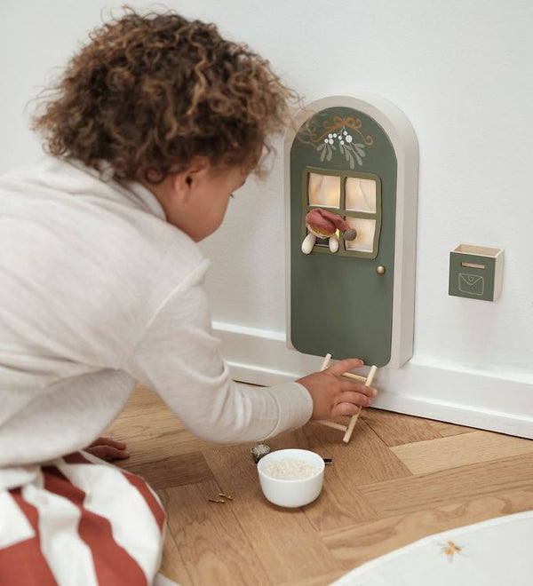 A child with curly hair kneels on the wooden floor, captivated by the Light-Up Elf Door on the wall. The miniature green door is adorned with a small wreath and ladder. Nearby, there's a tiny mailbox and a bowl filled with a white substance, creating an atmosphere that feels like stepping into another dimension.