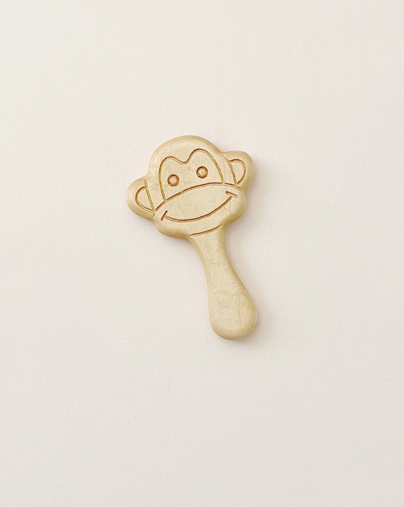 A Handmade Wooden Teether shaped like a monkey's head with a smiling face hangs against a plain white background. Crafted from cherry wood, the non-toxic baby rattle has wide eyes and a big curved smile etched into the simple design. The handle is smooth and elongated, perfect for little hands.