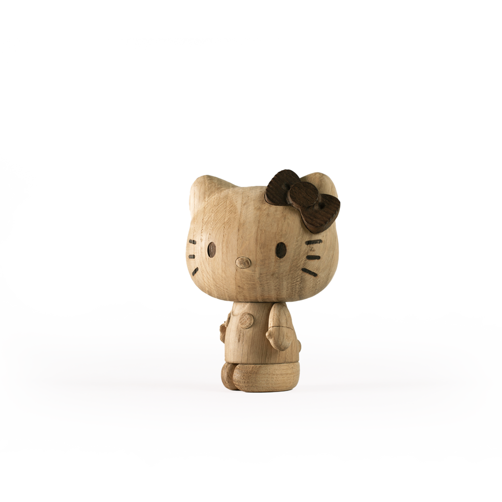 A wooden figurine resembling the Hello Kitty character is depicted. Crafted from European FSC White Oak, the figure features a large head with round eyes, a small nose, whiskers, and a distinctive bow on its left ear. The body is simple and rounded, aligning with contemporary design collectibles. The background is plain white. This product is named "Hello Kitty, Oak.