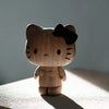 A wooden figurine of Hello Kitty, Oak, crafted from European FSC White Oak, stands on a surface, illuminated by a beam of light against a dark background. The contemporary design collectible features distinct black eyes, whiskers, and a bow on its left ear.