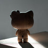 A wooden figurine with cat-like ears, inspired by Hello Kitty, stands on a flat surface illuminated by a beam of light coming from the side. Crafted from European FSC White Oak, its back casts a shadow on the surface, while the background remains dark and minimalistic.