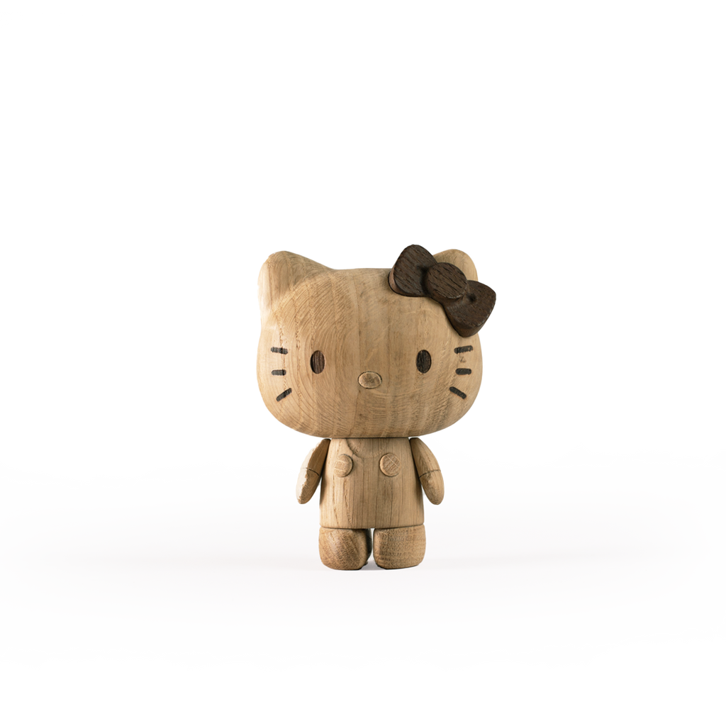 A wooden figurine resembling the fictional character Hello Kitty stands upright. Crafted from European FSC White Oak, it features carved detailing for the eyes, nose, and whiskers, along with a dark wooden bow on its left ear. This piece embodies Hello Kitty, Oak against a plain white background.