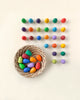 A basket of Grapat Mandala Rainbow Eggs, positioned next to neat rows of multicolored balls arranged in a grid on a light background.