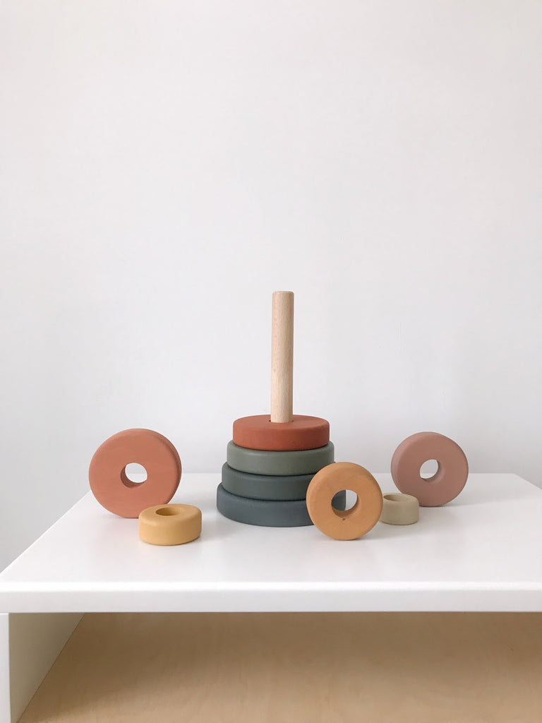 A simple, minimalist setup of a Wooden Pyramid Stacker - Green & Mustard with colored rings partially assembled on a dowel, placed on a white table against a pale background.