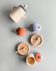 A flat lay of Raduga Grez Handmade Wooden Tea Set items on a grey backdrop. The handmade set includes a beige teapot with a black lid, three teacups in beige, red, and purple, two saucers with small spoons, and a lidded jar. The arrangement is minimalistic and neat.