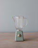 A Maileg Miniature Blender with a clear plastic jug and a light green base featuring a single round switch, perfect for children's toy pretend play. The Maileg Miniature Blender is placed on a light wooden surface with a pale blue background.